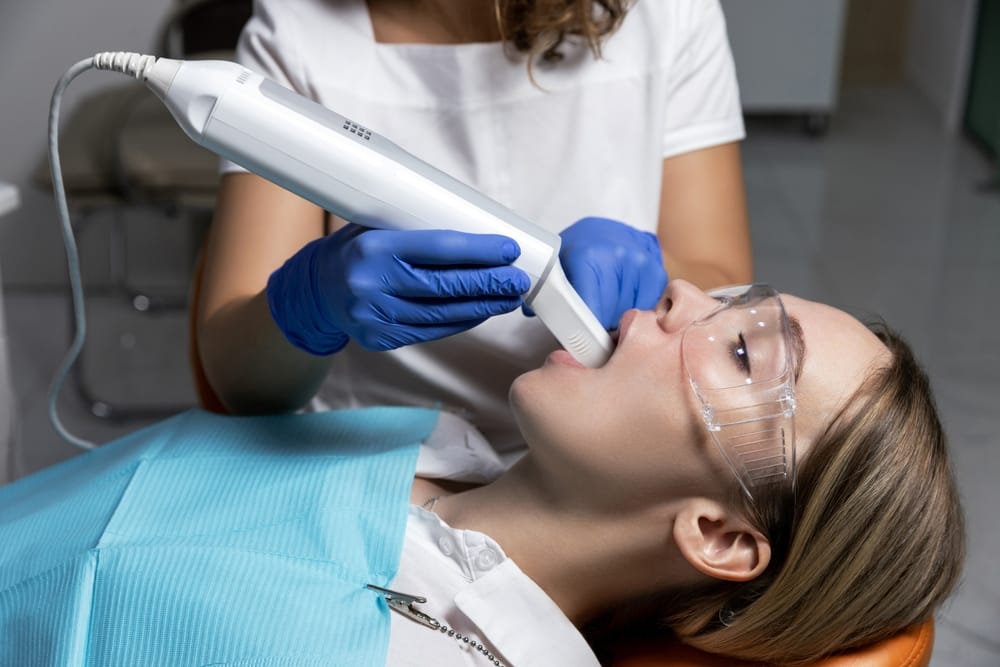 Dental Digital Scanner in Use – Smile Haven A dental professional uses a handheld digital scanner to capture images of a patient's teeth, with the patient wearing protective glasses and a blue bib – Smile Haven