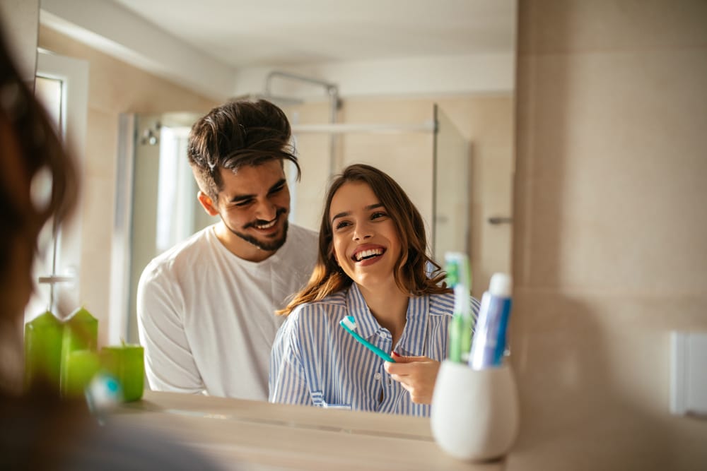 Couple Smiling in Bathroom – Smile Haven A cheerful couple stands together in a bathroom, smiling at their reflection in the mirror while the woman holds a toothbrush – Smile Haven