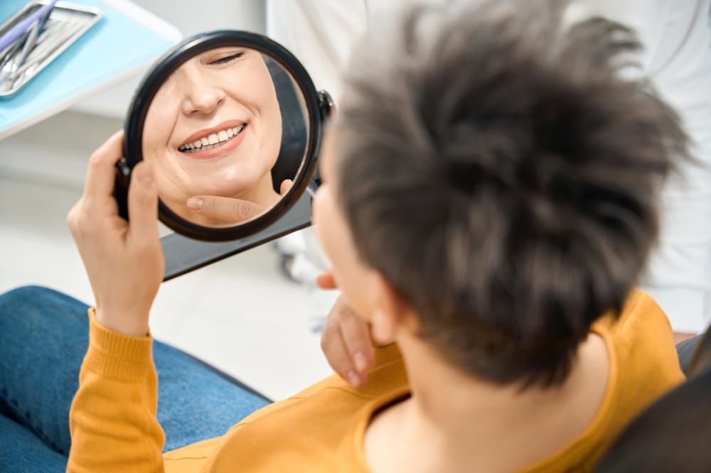 Patient Admiring Smile in Mirror – Smile Haven A person wearing a mustard yellow top smiles while looking at their reflection in a handheld mirror during a dental visit – Smile Haven