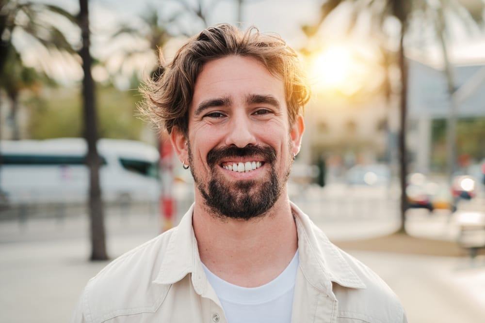 Smiling Man Outdoors – Smile Haven A cheerful man with wavy hair and a beard smiles while standing outside with palm trees and a sunset in the background – Smile Haven
