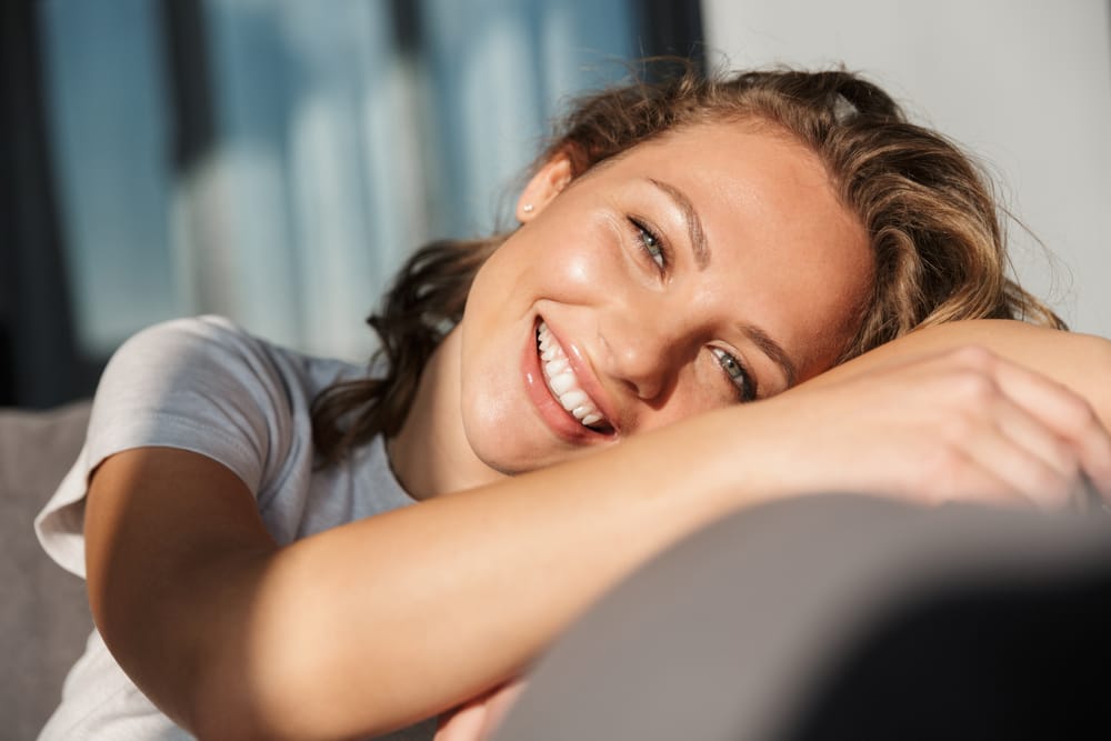 Smiling Woman Relaxing – Smile Haven A young woman with wavy blonde hair smiling warmly while resting her head on her arms, sitting indoors with soft natural light – Smile Haven