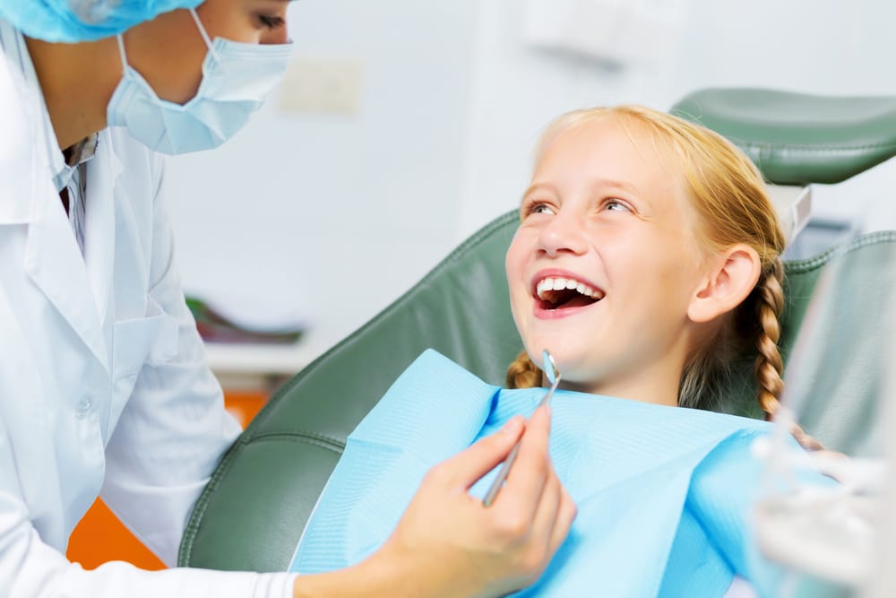 Child at Dental Appointment – Smile Haven A smiling young girl with blonde hair in braids sitting in a dental chair, wearing a blue bib, as a masked dentist holds a dental tool – Smile Haven