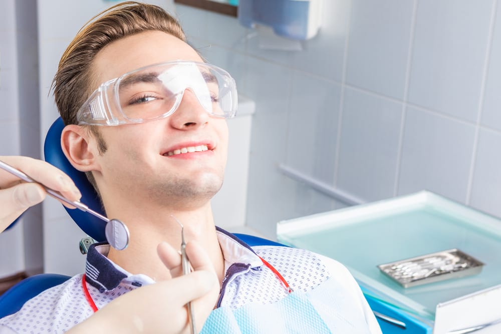 Young Man at Dentist – Smile Haven A young man wearing protective glasses smiles while sitting in a dental chair, as a dentist holds dental tools near his mouth – Smile Haven